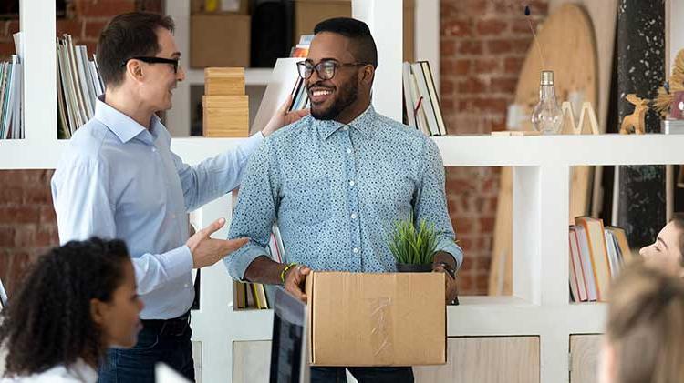 While standing in an office, a man is talking with another man about his 401(k) while carrying a box.